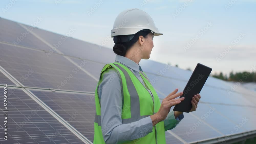 Beautiful caucasian female worker in signal vest and protective helmet stands near solar panels and checks condition of batteries using tablet. Beautiful caucasian female worker in signal vest and protective helmet stands near solar panels and checks condition of batteries using tablet.