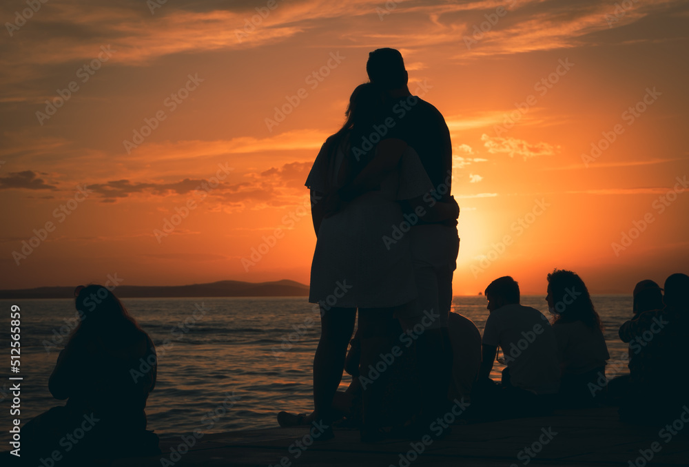 Zakochana para przytula się i patrzy na zachód słońca. Ludzie siedzący na promenadzie patrzący na zachód słońca. Zdjęcie pod światło,  zarys  sylwetek. - obrazy, fototapety, plakaty 