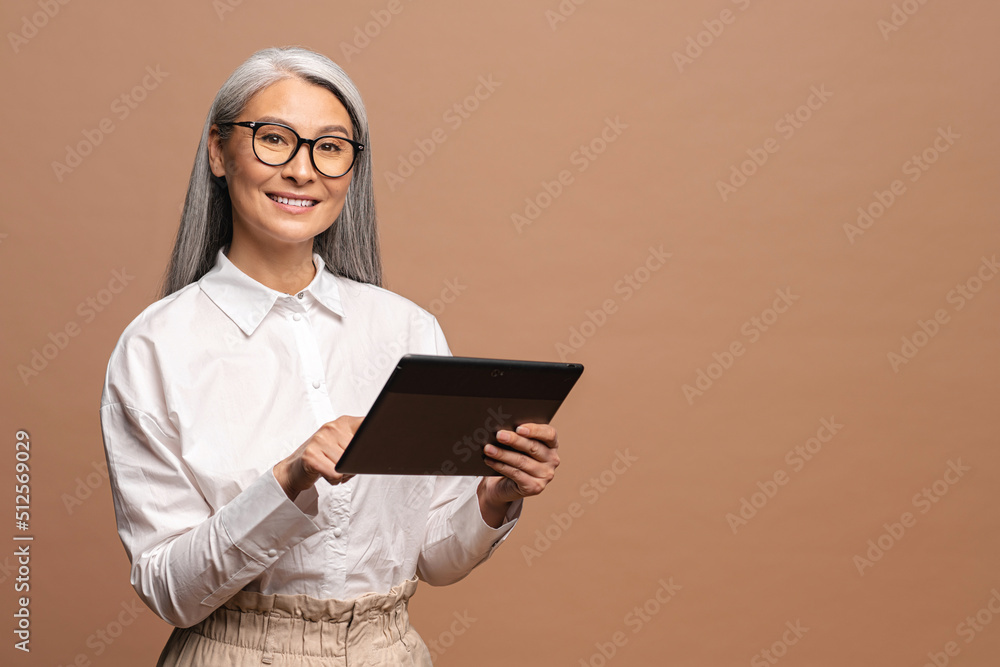 © Vadim Pastuh - Modern elderly senior woman in formal wear using digital tablet isolated on beige. Portrait of mature female office employee using online technology for doing business, computer app for accounting