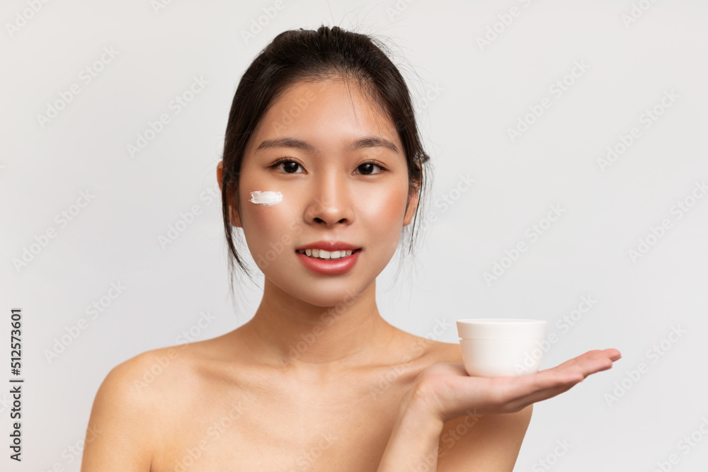Pretty korean woman with perfect skin applying moisturizing cream on her face and holding jar with blank space Pretty korean woman with perfect skin applying moisturizing cream on her face and holding jar with blank space