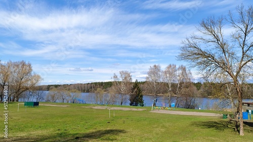 On the shore of the lake grow firs, birches, willows and other trees, there are cabins for changing clothes and created sports fields. On the opposite shore is a forest. The sky is blue with clouds