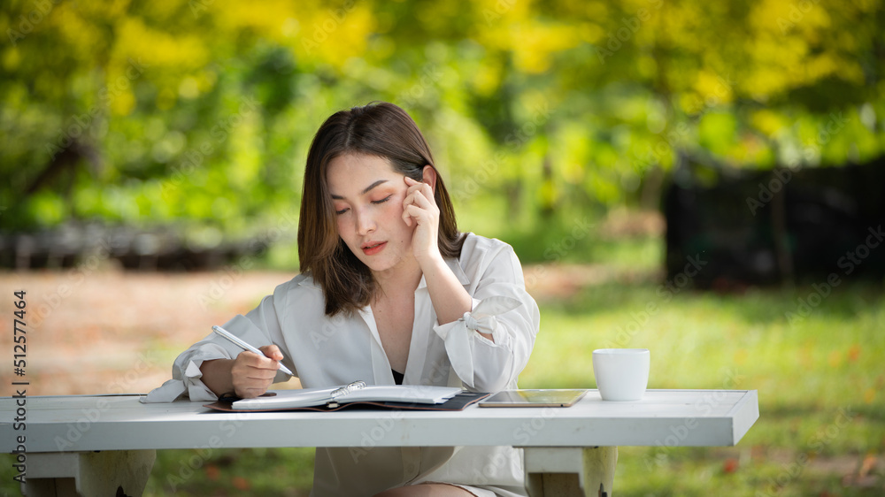 Thoughtful smile woman in park using notebook and writing, Portrait of a young charming business woman checking online Business work on her smart phone outdoors in the park on soft green back ground Thoughtful smile woman in park using notebook and writing, Portrait of a young charming business woman checking online Business work on her smart phone outdoors in the park on soft green back ground
