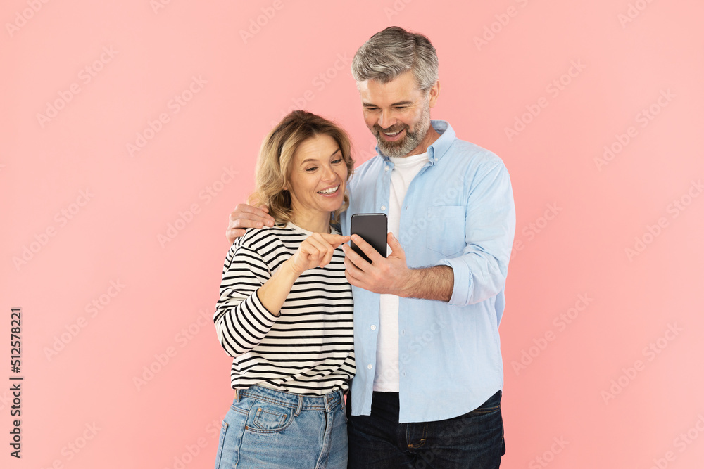 Cheerful Middle Aged Spouses Using Mobile Phone On Pink Background Cheerful Middle Aged Spouses Using Mobile Phone On Pink Background