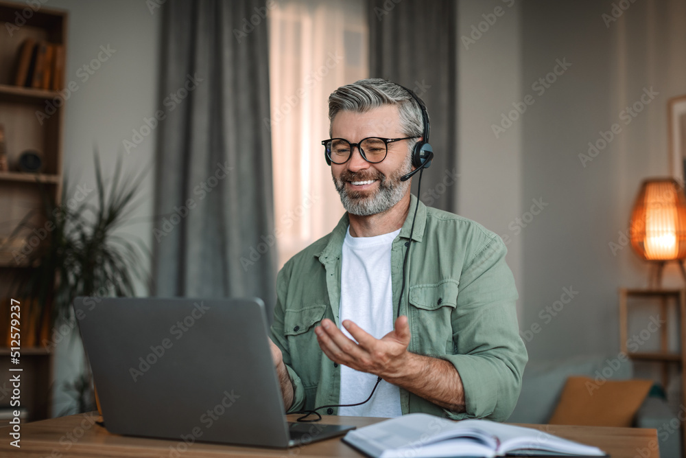 Happy senior european man with beard in headset and glasses gesticulates and looks at laptop Happy senior european man with beard in headset and glasses gesticulates and looks at laptop