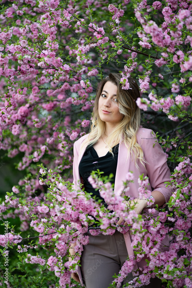 A very beautiful blonde in a pink jacket and a black blouse sensually poses standing among the flowers of cherry blossoms and smiling A very beautiful blonde in a pink jacket and a black blouse sensually poses standing among the flowers of cherry blossoms and smiling
