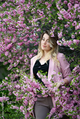 A very beautiful blonde in a pink jacket and a black blouse sensually poses standing among the flowers of cherry blossoms and smiling