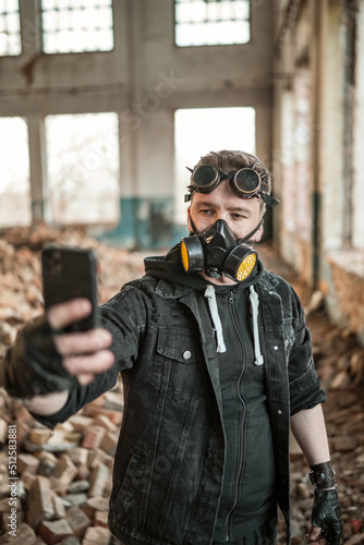 A brutal guy with an eyebrow piercing in cyberpunk or steampunk goggles on his forehead and a respirator on his face doing selfie on smartphone in ruined building. Apocalypse cyberpunk male portrait