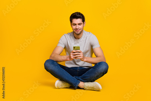Full body photo of nice brunet millennial guy sit look telephone wear t-shirt jeans footwear isolated on yellow background © deagreez