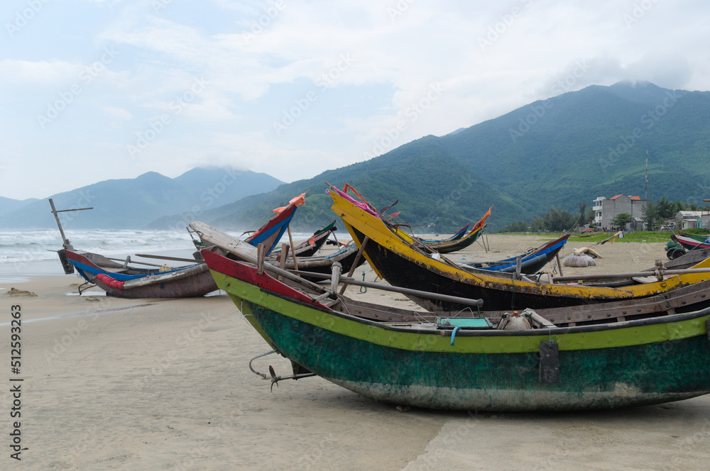 Fototapeta premium Curved fishing boats on coast of South China Sea, Lang Co, Vietnam. Sandy beach with small Vietnamese fishing boats. Green mountains, sea waves and slightly foggy from water splashes in background
