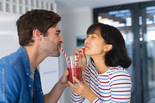 Happy multiracial young couple drinking fruits smoothie through straw in kitchen