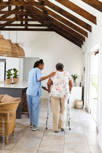 African american female doctor assisting senior woman in walking with walker on floor at home photo