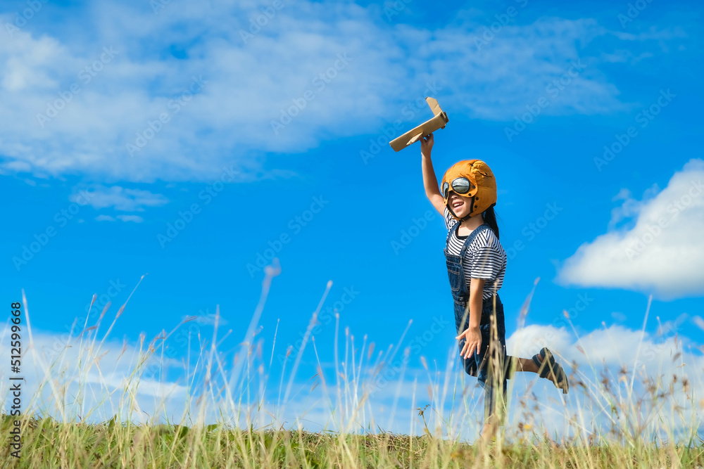 Cute little girl running through the meadow on a sunny day with a toy plane in hand. Happy kid playing with cardboard plane against blue summer sky background. Childhood dream imagination concept. Cute little girl running through the meadow on a sunny day with a toy plane in hand. Happy kid playing with cardboard plane against blue summer sky background. Childhood dream imagination concept.