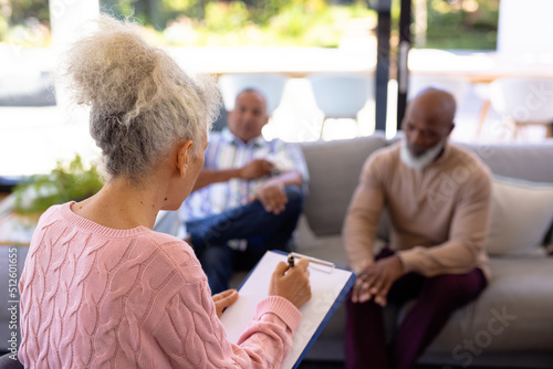 Multiracial senior woman with clipboard listening to men sharing their problems in retirement home