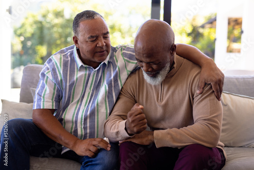 Multiracial senior man consoling male friend crying while sitting on sofa in nursing home