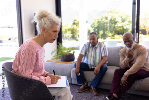 Fotografie, Tablou Multiracial female therapist with clipboard guiding seniors in group therapy ses