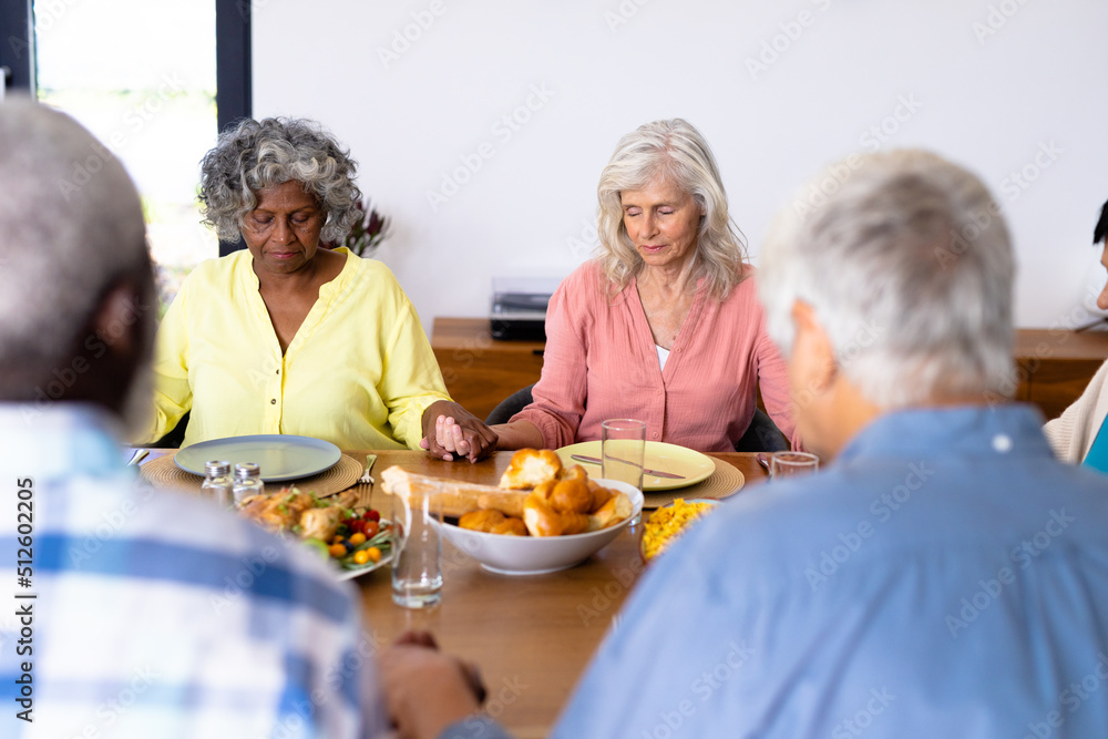 Multiracial senior friends holding hands and saying grace at dining table in nursing home