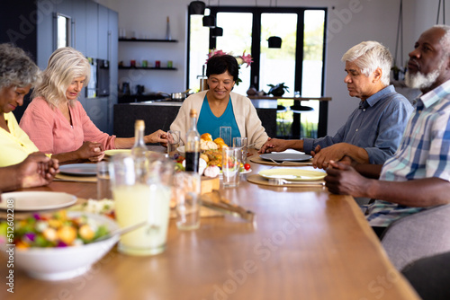 Multiracial senior friends with food on dining table holding hands and saying grace in nursing home