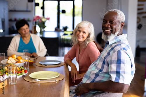 Portrait of multiracial senior friends with plates and food on dining table sitting in nursing home