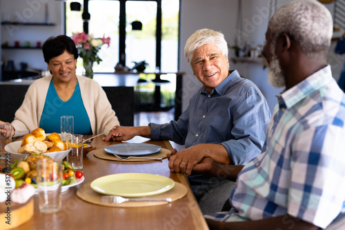 Multiracial smiling senior friends holding hands and talking at dining table in nursing home