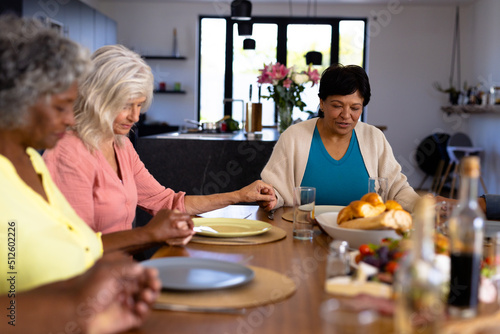 Multiracial senior female friends holding hands and saying grace at dining table in nursing home