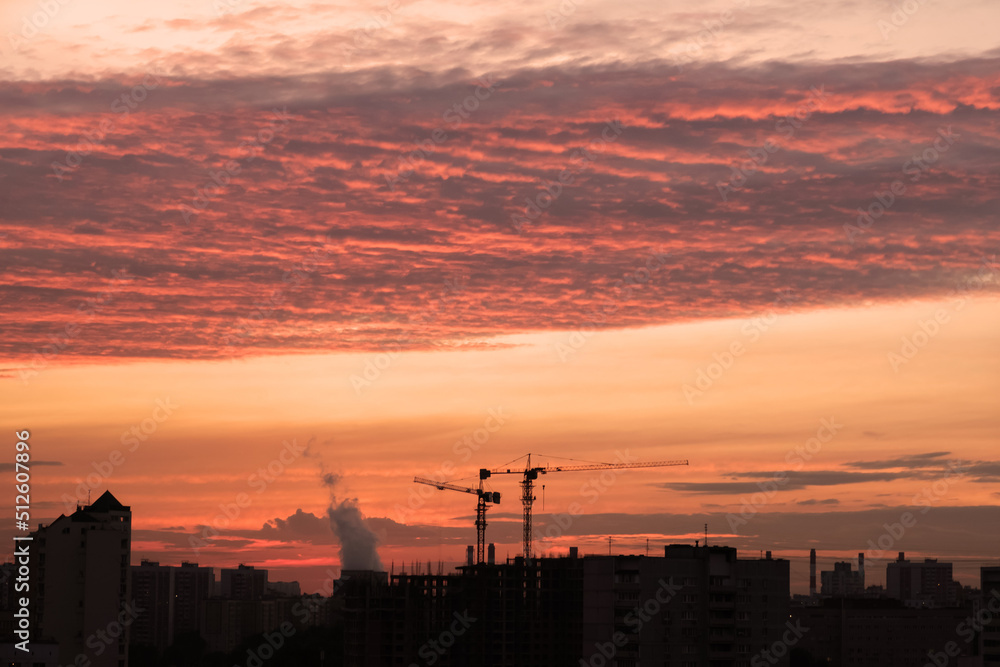 Industrial landscape of the sunset sky. Silhouettes of multi-storey residential buildings, construction cranes and pipes of factories on red-blue sunset background. Sun illuminates cirrocumulus clouds