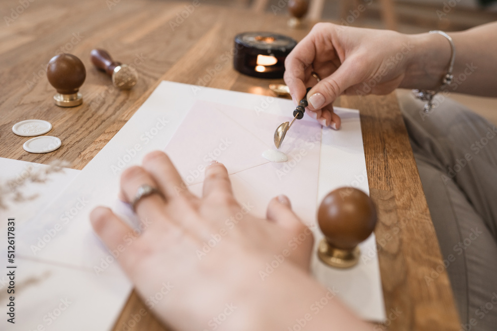 Obraz premium Close-up of hands of unrecognizable woman pouring liquid white wax seal with spoon on pink greeting envelope on desk.