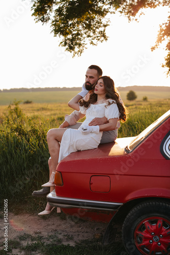 A happy couple in love sits and hugs on the trunk of a red retro car at sunset.