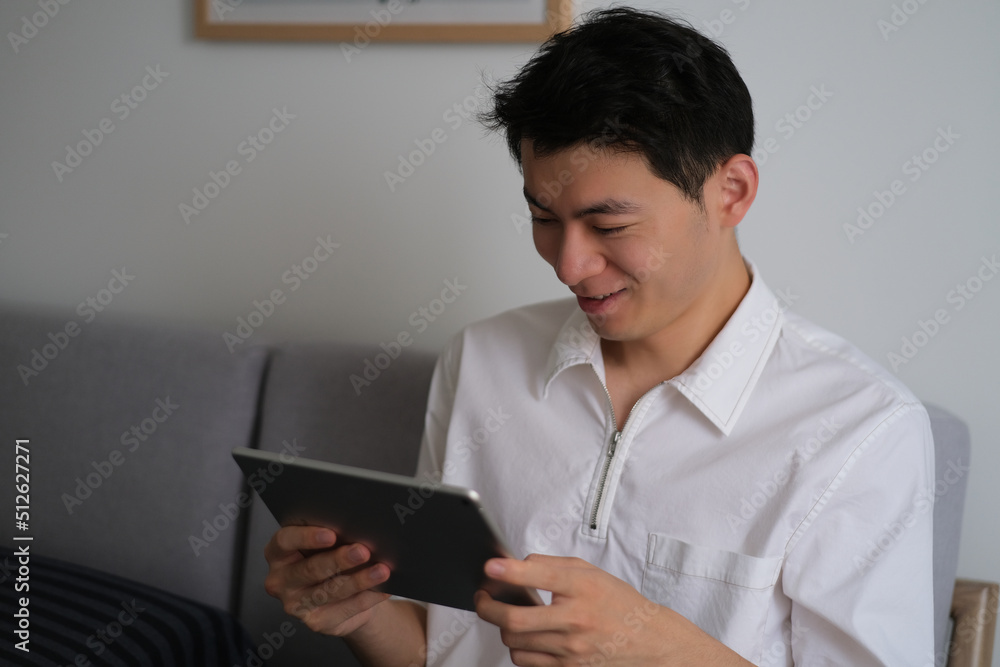 Handsome Asian young man looking at tablet computer with smiling at home Handsome Asian young man looking at tablet computer with smiling at home