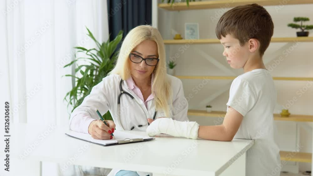 Smiling senior female family doctor in uniform consulting little kid patient with a broken hand at checkup meeting in clinic Smiling senior female family doctor in uniform consulting little kid patient with a broken hand at checkup meeting in clinic