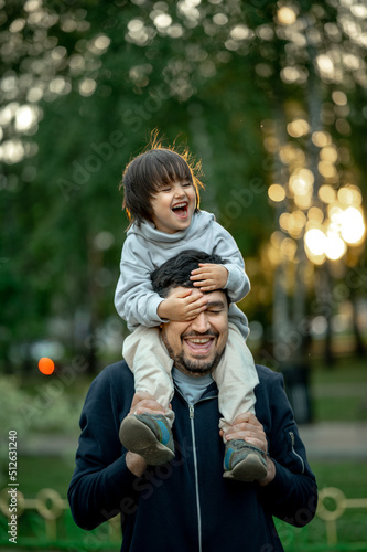 a cute three-year-old boy sits on dad's shoulders, dad walks with his son on a summer evening in the park, a happy family, a father spends time with his child