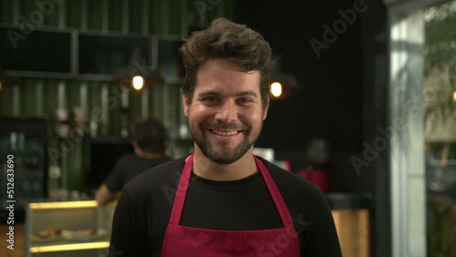 Portrait of young barista standing inside cafe smiling. Handsome employee worker staff wearing apron