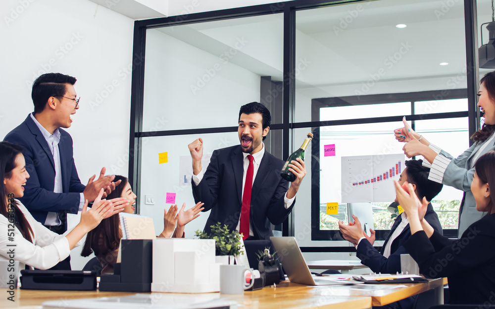 Caucasian handsome businessman wearing formal suit, holding bottle of wine to celebrate his success. Asian smiley colleagues clap hands and sitting in meeting room at indoor office or workplace. Caucasian handsome businessman wearing formal suit, holding bottle of wine to celebrate his success. Asian smiley colleagues clap hands and sitting in meeting room at indoor office or workplace.