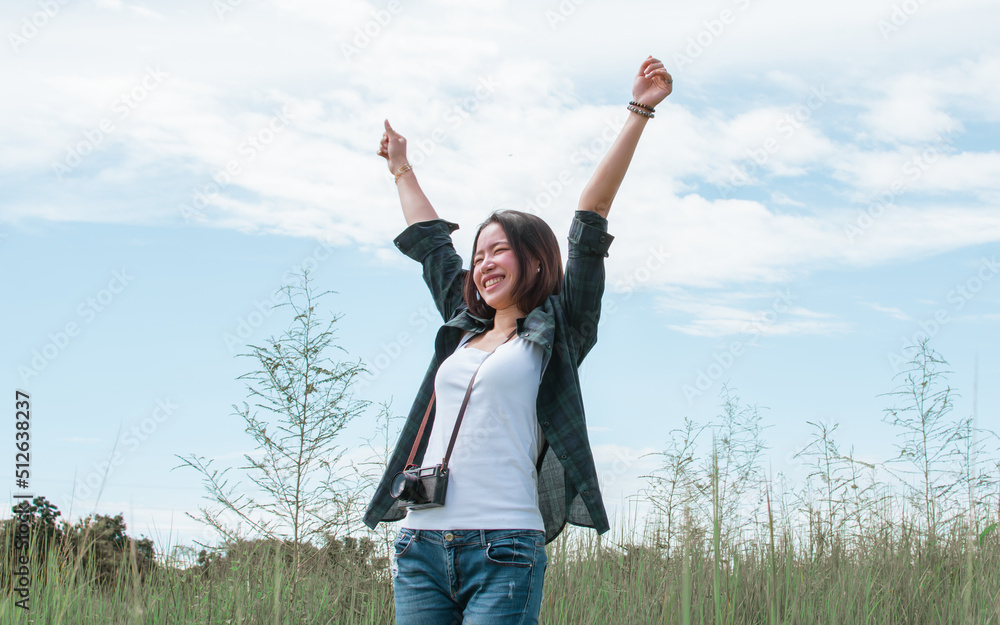 Beautiful Asian female tourist wearing casual clothes and jeans, smiling, raising hand with happiness and relaxation, traveling alone on summer vacation holiday with background of green field and sky. Beautiful Asian female tourist wearing casual clothes and jeans, smiling, raising hand with happiness and relaxation, traveling alone on summer vacation holiday with background of green field and sky.