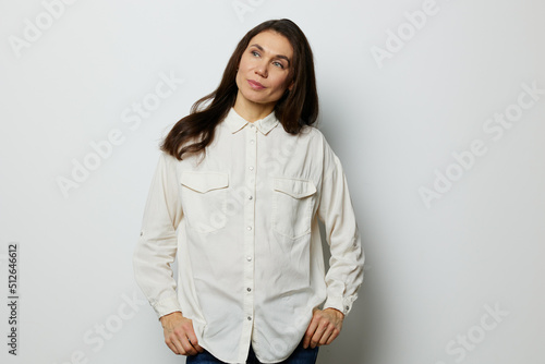 a beautiful, attractive, adult woman, stands with black hair flowing in the wind in a white shirt and holds her hand behind her head. Studio photo with an empty space on the background