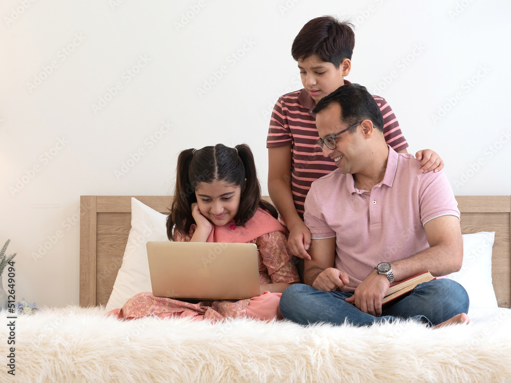 Indian father with two teenage children sitting on the bed together at home using laptop for movie, homework, online learning. Happy family, single father , education or father's day concept. Indian father with two teenage children sitting on the bed together at home using laptop for movie, homework, online learning. Happy family, single father , education or father's day concept.
