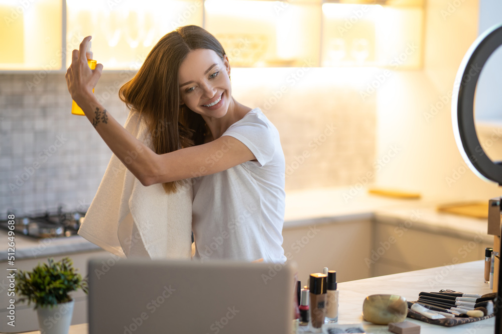 Woman holding spray near hair looking at laptop