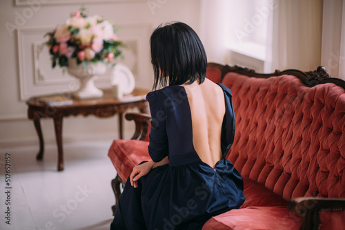 Rear view of a brunette woman in sexy blue dress with bare back sitting on sofa.