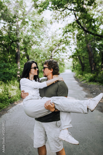 A bearded long-haired stylish man holds his beloved hippie brunette woman in sunglasses in his arms and circles her in nature in the park. Portrait, photo of newlyweds, concept.