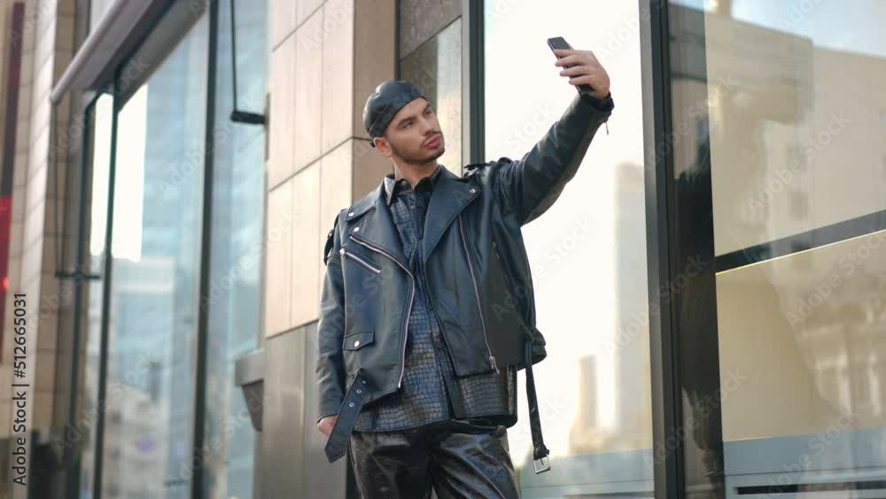 Portrait of confident Caucasian gay man taking selfie on smartphone standing on city street outdoors. Proud young LGBTQ person enjoying weekend leisure outdoors. Style and confidence concept Portrait of confident Caucasian gay man taking selfie on smartphone standing on city street outdoors. Proud young LGBTQ person enjoying weekend leisure outdoors. Style and confidence concept