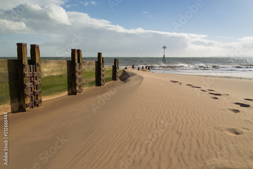 A beach scene with a sandbank  containing footprints leaning against a wooden groyne that leads to the sea and a lane marker on the horizon as waves hit the shoreline. © Mark