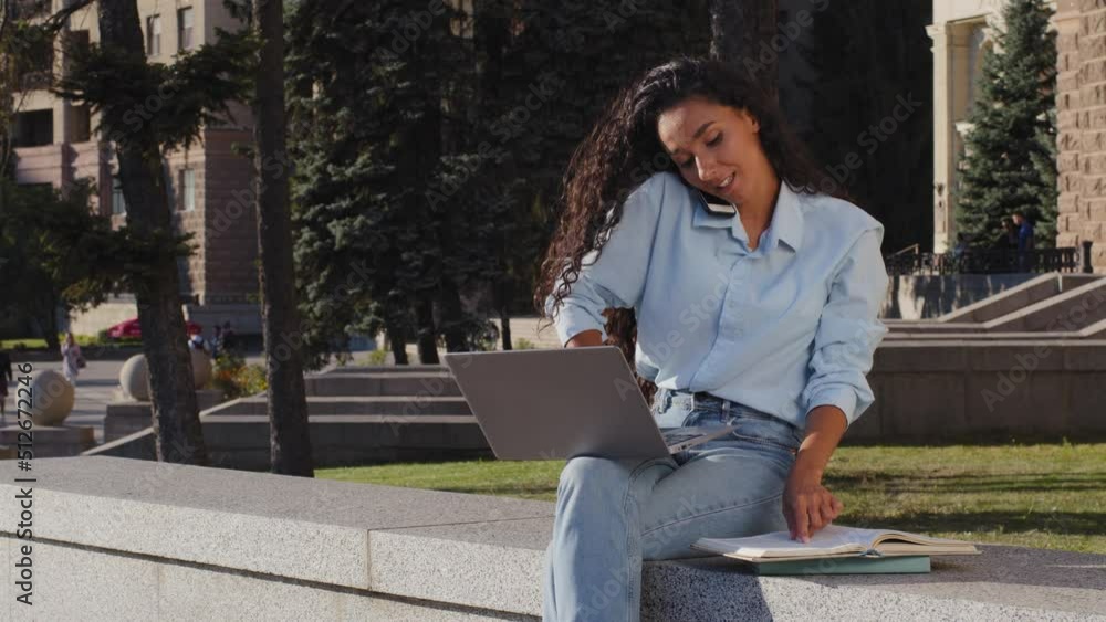 Multitasking young girl business woman freelancer study working with laptop talking mobile phone reading book making notes answering call writes sitting on street office college building background Multitasking young girl business woman freelancer study working with laptop talking mobile phone reading book making notes answering call writes sitting on street office college building background