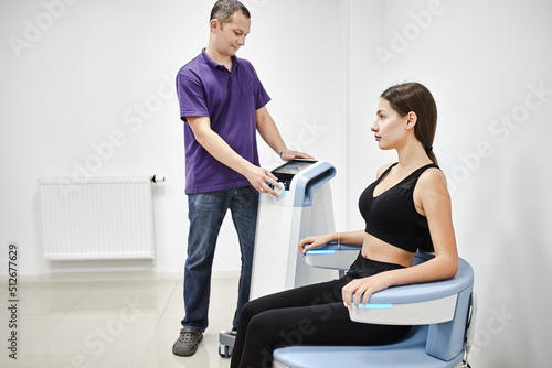 Young woman sitting on electromagnetic chair for stimulation of deep pelvic floor muscles and restoring neuromuscular control at the clinic while doctor control process