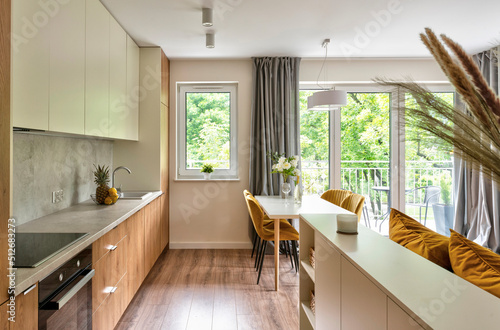 Modern interior of kitchen with wooden furniture and counter and cabinet. Open space with dining room with table and mustard chairs and big window. New apartment at home. © photosbysabkapl