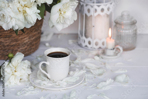 A beautiful postcard. A white coffee cup with a saucer, candles and a vase with a bouquet of white peonies. Beautiful still life. Spring, summer time. The concept of "Good morning".