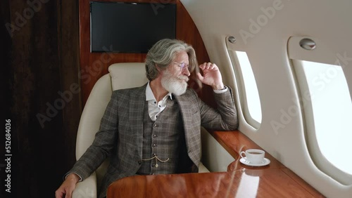 Stylish mature millionaire man wearing classical suit luxury flying in private airplane. Confident CEO businessman looking through corporate jet window. Luxury traveling in private jet. photo