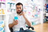 Disabled man in wheelchair talking on phone while choosing medicine in pharmacy.