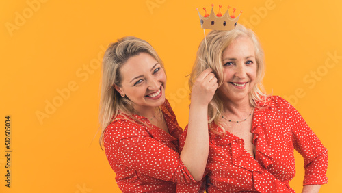 Medium studio shot on yellow background of two european blonde-haired women in the 40s wearing similar outfits. One woman holding a prop crown over the head of the other woman. High quality photo