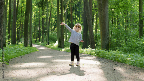 cheerful child in striped t-shirt waves her hand at camera on summer sunny day in park among tall green trees. girl is happy and shows her joy.