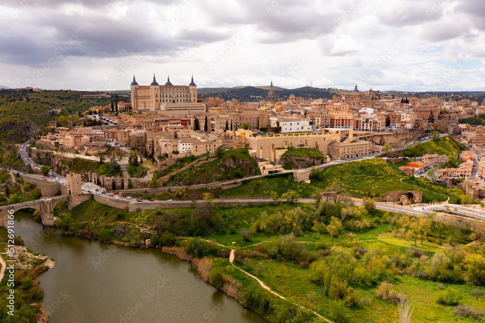Picturesque aerial view of Toledo cityscape on green hilly banks of Tagus river in early spring overlooking fortified Alcazar castle framed by four towers, Spain