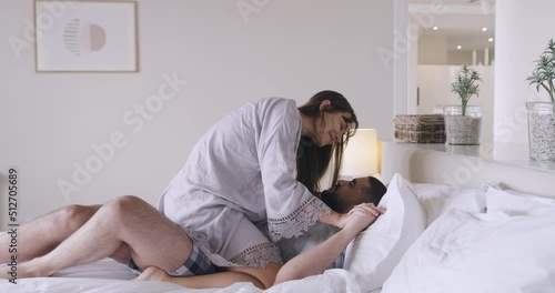 Loving playful couple lying in bed kissing each other good morning and playing in their pajamas. Happy husband lying while the wife sits on top of him cuddling in bed together in their modern bedroom